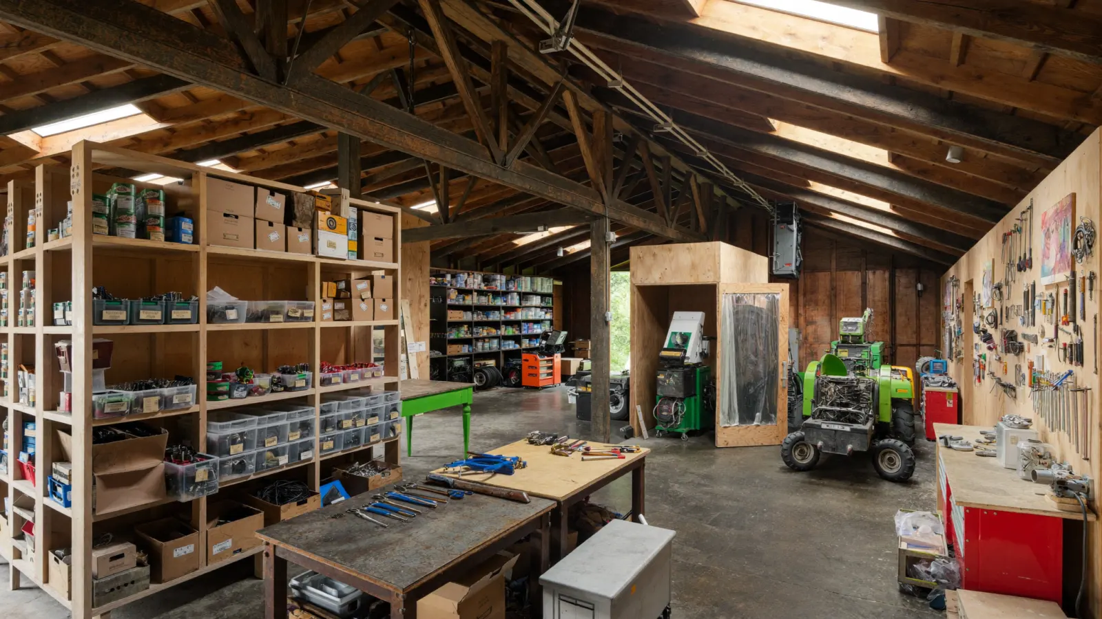 A large community workshop with timber trusses, shelves of labelled spare parts, workbenches, tools and a small tractor under repair